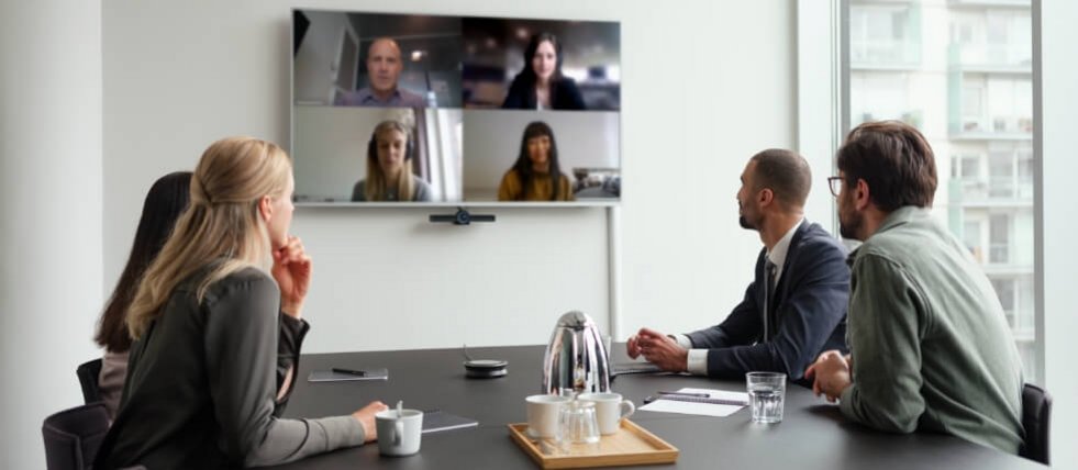 Eine Gruppe von Mitarbeitenden sitzt im Konferenzraum und schaut auf einen Monitor, wo andere Personen per Video zugeschaltet sind