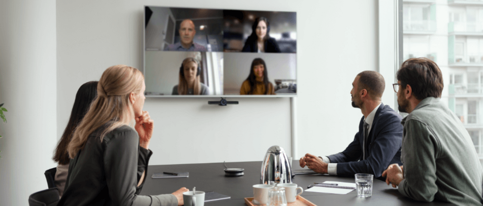 Vier Personen sitzen an einem Tisch und blicken auf einen großen Monitor auf dem weitere Personen zu sehen sind, die an einer Videokonferenz teilnehmen.