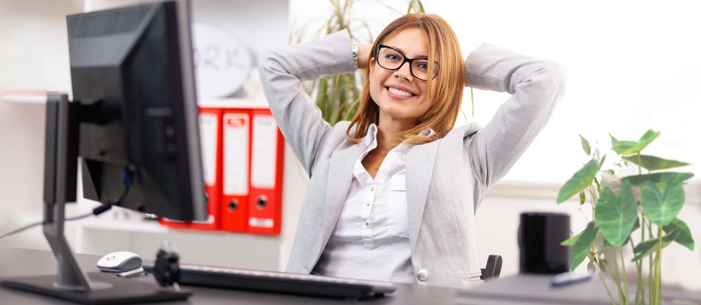Lächelnde Frau sitzt lächelnd und entspannt mit hinter dem Kopf verschränkten Armen vor einem Computer.